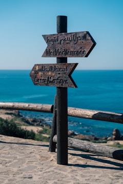 A wooden signpost with two arrows points towards the Mediterranean Sea and the Atlantic Ocean. The backdrop features a bright blue sky and a clear view of an expansive body of water. The ground is sandy with some rocks and sparse vegetation.