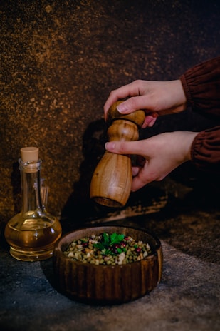 A person uses a wooden pepper grinder over a rustic bowl filled with a colorful mixture of grains and herbs. A glass bottle containing liquid, possibly oil or vinegar, is positioned nearby on a textured surface.