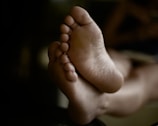 The image depicts a close-up view of a person's bare feet. The focus is on the soles and toes, with a soft, blurred background. The lighting is low and emphasizes the natural texture of the skin.