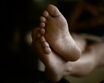 The image depicts a close-up view of a person's bare feet. The focus is on the soles and toes, with a soft, blurred background. The lighting is low and emphasizes the natural texture of the skin.