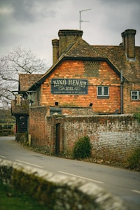 A rustic brick building features a sign reading 'King Henry VIII Country Pub & Kitchen'. The structure has a classic, old-fashioned appearance with chimneys and a satellite antenna on the roof. The scene includes a narrow road lined with a moss-covered brick wall and some shrubs, with bare trees visible in the background.