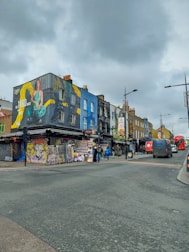 A bustling street scene featuring colorful buildings with vibrant murals and graffiti. The area is lively, with several people walking along the sidewalk, and various storefronts, including a tattoo and piercing shop. Bright, artistic designs and a large advertisement on the building create an eclectic atmosphere. The sky is cloudy, adding a subdued tone to the overall vibe of the street.