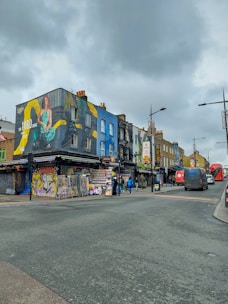 A bustling street scene featuring colorful buildings with vibrant murals and graffiti. The area is lively, with several people walking along the sidewalk, and various storefronts, including a tattoo and piercing shop. Bright, artistic designs and a large advertisement on the building create an eclectic atmosphere. The sky is cloudy, adding a subdued tone to the overall vibe of the street.