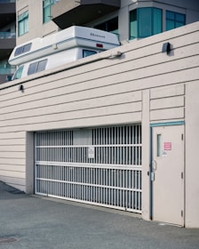 A white vehicle with 'Okanagan' branding is partially visible above a large white metal gate of a parking structure adjacent to a modern building. The building features large windows and multiple balconies. The scene has an urban aesthetic.