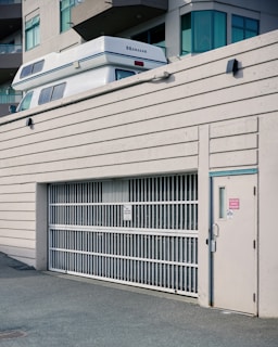 A white vehicle with 'Okanagan' branding is partially visible above a large white metal gate of a parking structure adjacent to a modern building. The building features large windows and multiple balconies. The scene has an urban aesthetic.