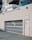 A white vehicle with 'Okanagan' branding is partially visible above a large white metal gate of a parking structure adjacent to a modern building. The building features large windows and multiple balconies. The scene has an urban aesthetic.