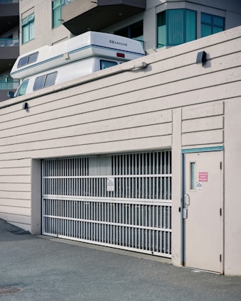 A white vehicle with 'Okanagan' branding is partially visible above a large white metal gate of a parking structure adjacent to a modern building. The building features large windows and multiple balconies. The scene has an urban aesthetic.