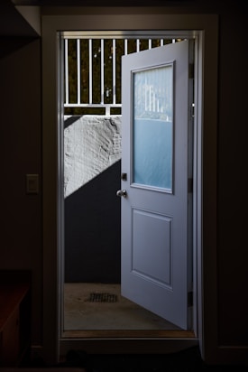 An open white door leading to a partially lit outdoor space with a concrete floor and a wall topped by a metal railing. Shadows cast on the wall create a contrast between light and dark areas.