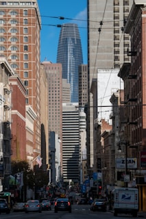 A bustling urban street scene featuring tall skyscrapers, with a prominent blue-glass building in the background. The street is lined with vehicles and pedestrians, creating a sense of city life. Overhead wires are visible, and various signs and advertisements can be seen along the buildings. The sky is clear with a vibrant blue color.