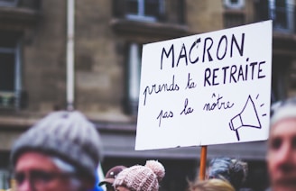 A protest scene with a focus on a handwritten sign in French that reads 'MACRON prends ta RETRAITE pas la nôtre' with a drawing of a megaphone. People in winter clothes and blurry background buildings are visible.