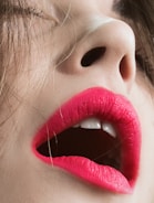 A close-up image of a person's face, focusing on the mouth and nose. The lips are adorned with vivid pink lipstick and slightly parted, revealing white teeth. The skin tone is fair, and strands of brown hair partially cover the face.