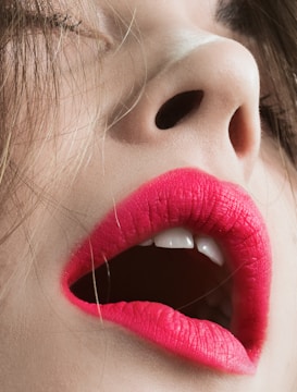 A close-up image of a person's face, focusing on the mouth and nose. The lips are adorned with vivid pink lipstick and slightly parted, revealing white teeth. The skin tone is fair, and strands of brown hair partially cover the face.
