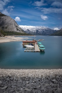 A serene lake surrounded by majestic snow-capped mountains, with a wooden dock extending into the water. Several boats are moored at the dock, with a turquoise tour boat prominently visible. The sky is partly cloudy, and the shoreline is rocky with patches of greenery.