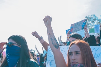 A group of individuals is gathered at a protest, with many holding signs. A prominent figure in the foreground raises a fist, symbolizing solidarity or defiance. The atmosphere is one of activism and unity, with people wearing bandanas and sunglasses.