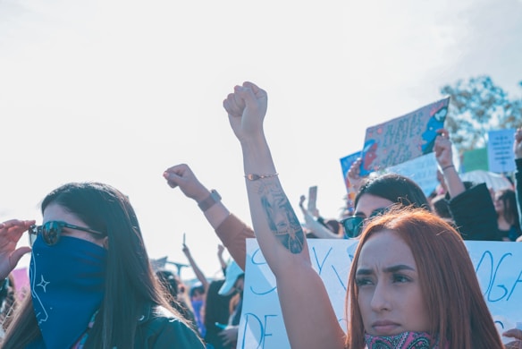 A group of individuals is gathered at a protest, with many holding signs. A prominent figure in the foreground raises a fist, symbolizing solidarity or defiance. The atmosphere is one of activism and unity, with people wearing bandanas and sunglasses.