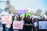 A group of people are gathered holding various protest signs with feminist messages. The scene takes place outdoors, with trees and a clear sky in the background. Some people are wearing scarves, hats, and sunglasses.