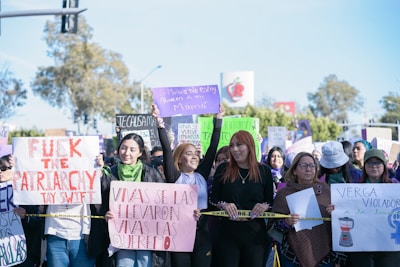 A group of people are gathered holding various protest signs with feminist messages. The scene takes place outdoors, with trees and a clear sky in the background. Some people are wearing scarves, hats, and sunglasses.