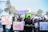 A group of people are gathered holding various protest signs with feminist messages. The scene takes place outdoors, with trees and a clear sky in the background. Some people are wearing scarves, hats, and sunglasses.