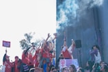 A group of people wearing red gather together holding signs and raising their hands in the air. Some are holding up items like drums and microphones. Smoke or mist is visible in the air, blending with a backdrop of a building and blue sky. The signs have symbols and messages related to feminism and activism.