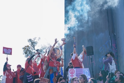 A group of people wearing red gather together holding signs and raising their hands in the air. Some are holding up items like drums and microphones. Smoke or mist is visible in the air, blending with a backdrop of a building and blue sky. The signs have symbols and messages related to feminism and activism.