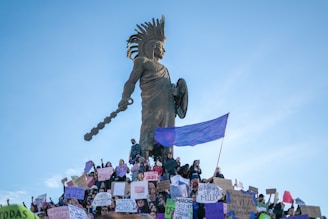 A large gathering of people holding signs and banners at the base of a tall statue which depicts a warrior figure. The statue has distinct features such as a headdress and a raised club. A purple flag is prominently displayed among the crowd.