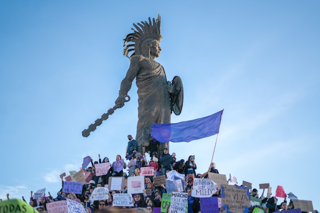 A large gathering of people holding signs and banners at the base of a tall statue which depicts a warrior figure. The statue has distinct features such as a headdress and a raised club. A purple flag is prominently displayed among the crowd.