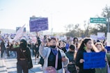 A large group of people are participating in a march or protest. They are holding various signs with messages, some of them in Spanish. The participants are diverse, wearing casual clothing, some with face coverings. The atmosphere is energized and focused. In the background, there are trees and a street sign indicating directions.