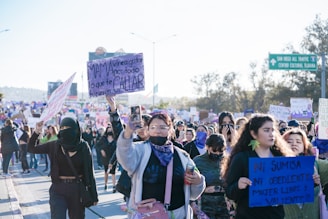 A large group of people are participating in a march or protest. They are holding various signs with messages, some of them in Spanish. The participants are diverse, wearing casual clothing, some with face coverings. The atmosphere is energized and focused. In the background, there are trees and a street sign indicating directions.