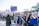 A large group of people are participating in a march or protest. They are holding various signs with messages, some of them in Spanish. The participants are diverse, wearing casual clothing, some with face coverings. The atmosphere is energized and focused. In the background, there are trees and a street sign indicating directions.