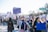 A large group of people are participating in a march or protest. They are holding various signs with messages, some of them in Spanish. The participants are diverse, wearing casual clothing, some with face coverings. The atmosphere is energized and focused. In the background, there are trees and a street sign indicating directions.