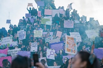 A large group of people, mostly women, gather for a protest or demonstration. Many are holding signs with slogans and messages advocating for rights and against violence. The setting appears to be outdoors with some haze or smoke visible in the air.