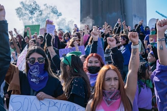 A large group of people gathered together, many with their fists raised in the air. Most are wearing colorful bandanas and clothing, and holding banners or signs. The atmosphere suggests a protest or rally. The crowd appears energetic and united.