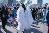 A crowded outdoor scene featuring a group of people participating in a public gathering or protest. The focus is on a person wearing large angel wings made of white material. Surrounding this individual, participants hold signs with various messages. The atmosphere suggests activism and collective energy. Buildings and trees are visible in the background under the clear sky.