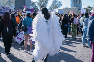 A crowded outdoor scene featuring a group of people participating in a public gathering or protest. The focus is on a person wearing large angel wings made of white material. Surrounding this individual, participants hold signs with various messages. The atmosphere suggests activism and collective energy. Buildings and trees are visible in the background under the clear sky.
