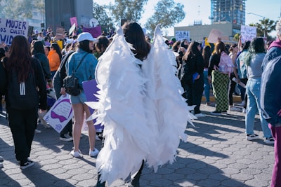 A crowded outdoor scene featuring a group of people participating in a public gathering or protest. The focus is on a person wearing large angel wings made of white material. Surrounding this individual, participants hold signs with various messages. The atmosphere suggests activism and collective energy. Buildings and trees are visible in the background under the clear sky.