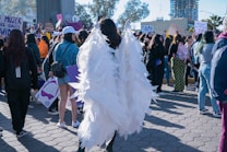 A crowded outdoor scene featuring a group of people participating in a public gathering or protest. The focus is on a person wearing large angel wings made of white material. Surrounding this individual, participants hold signs with various messages. The atmosphere suggests activism and collective energy. Buildings and trees are visible in the background under the clear sky.