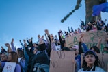 A group of people gathered outdoors, raising their fists in solidarity, with some holding signs with writings and symbols related to feminism and social justice. The crowd is diverse, with many individuals wearing masks and bandanas. The sky is clear and blue, creating a vivid backdrop.
