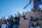 A group of people gathered outdoors, raising their fists in solidarity, with some holding signs with writings and symbols related to feminism and social justice. The crowd is diverse, with many individuals wearing masks and bandanas. The sky is clear and blue, creating a vivid backdrop.