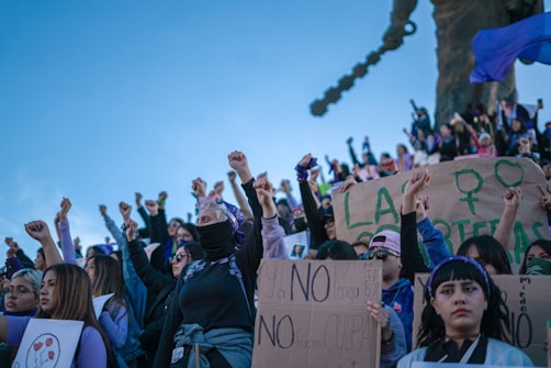 A group of people gathered outdoors, raising their fists in solidarity, with some holding signs with writings and symbols related to feminism and social justice. The crowd is diverse, with many individuals wearing masks and bandanas. The sky is clear and blue, creating a vivid backdrop.