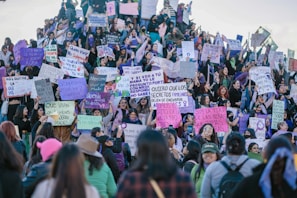A large group of people, mostly women, are gathered together holding signs with various messages and slogans. The signs are written in Spanish and appear to be advocating for women’s rights or protesting against gender-based violence. The crowd is diverse, with attendees of different ages and expressions, and many have serious or determined looks. The setting appears to be outdoors, possibly on a flight of steps or a sloped surface.