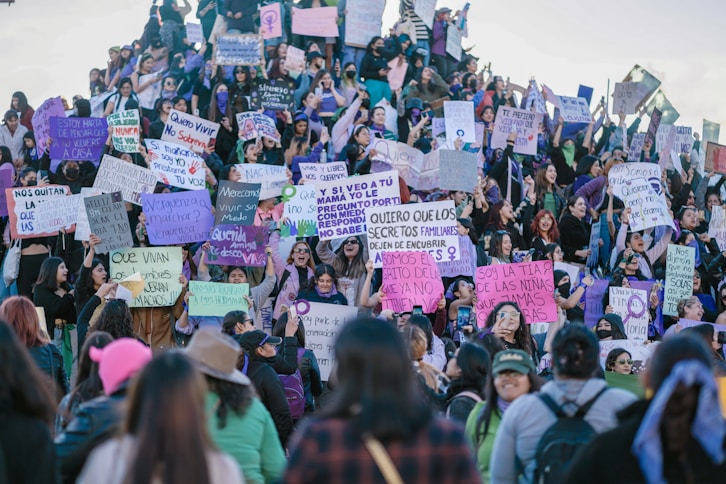 A large group of people, mostly women, are gathered together holding signs with various messages and slogans. The signs are written in Spanish and appear to be advocating for women’s rights or protesting against gender-based violence. The crowd is diverse, with attendees of different ages and expressions, and many have serious or determined looks. The setting appears to be outdoors, possibly on a flight of steps or a sloped surface.