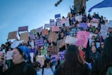 A large group of people gathering for a protest or demonstration, many holding signs with messages in Spanish advocating for women's rights and safety. The group appears to be standing on a staircase or slope with some people elevated higher than others. The sky is clear and blue, and the participants display various expressions of determination and solidarity.