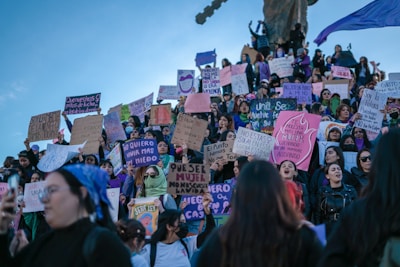 A large group of people gathering for a protest or demonstration, many holding signs with messages in Spanish advocating for women's rights and safety. The group appears to be standing on a staircase or slope with some people elevated higher than others. The sky is clear and blue, and the participants display various expressions of determination and solidarity.