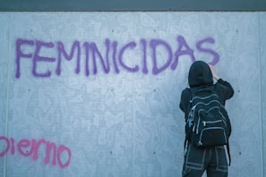 A person wearing a black hoodie and backpack is spray painting the word 'FEMINICIDAS' in bold purple letters on a grey concrete wall. The person appears to be focused on their task. Additional graffiti in pink can be seen partially at the bottom left.