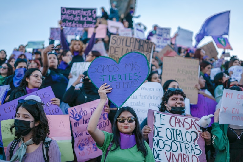 A large group of people gathered in a public demonstration, holding signs with messages related to empowerment and protest. The crowd is diverse, with individuals displaying expressions of determination and solidarity. Many participants are wearing purple clothing or accessories, and one prominently holds a heart-shaped sign.