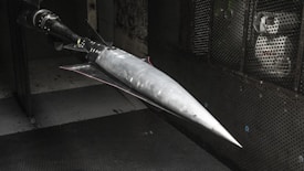A model of a sleek, pointed aerospace vehicle with a sharp nose and aerodynamic fins is mounted inside a testing environment. The surroundings are metallic and industrial, with a perforated surface and visible instrumentation.
