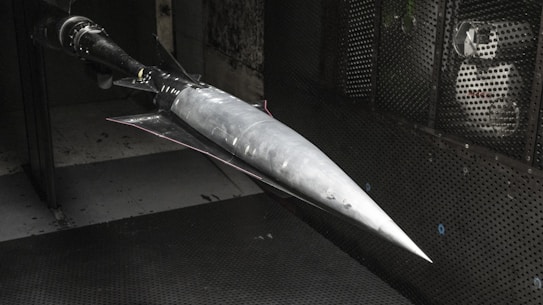 A model of a sleek, pointed aerospace vehicle with a sharp nose and aerodynamic fins is mounted inside a testing environment. The surroundings are metallic and industrial, with a perforated surface and visible instrumentation.