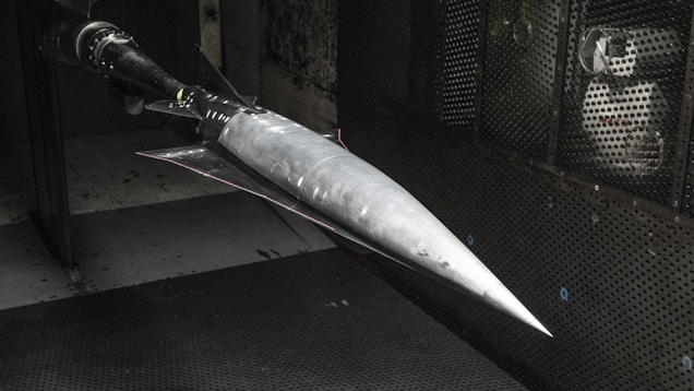 A model of a sleek, pointed aerospace vehicle with a sharp nose and aerodynamic fins is mounted inside a testing environment. The surroundings are metallic and industrial, with a perforated surface and visible instrumentation.