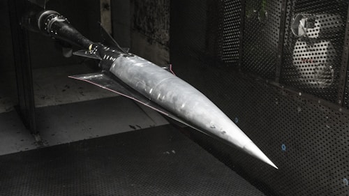 A model of a sleek, pointed aerospace vehicle with a sharp nose and aerodynamic fins is mounted inside a testing environment. The surroundings are metallic and industrial, with a perforated surface and visible instrumentation.