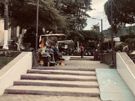 A park scene featuring several people near a vendor stall under trees. Stairs lead up to the area from the foreground, and there are pathways marked with accessibility symbols. The park is surrounded by greenery and has a tranquil atmosphere.